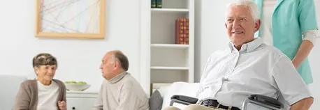 An elderly man in a wheelchair smiles in the foreground, while a caregiver stands beside him. In the background, an older couple sits on a couch, engaged in conversation. The room is bright and decorated with a painting and books on a shelf.