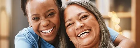 Two women are smiling joyfully. One woman, wearing a blue shirt, has short hair. The other woman has long, gray hair. They are close together, suggesting a warm, friendly relationship. The background is softly blurred with warm colors.