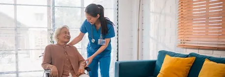 A nurse in blue scrubs smiles while helping an elderly person seated in a wheelchair. They are in a cozy room with large windows, a teal sofa, and yellow cushions.
