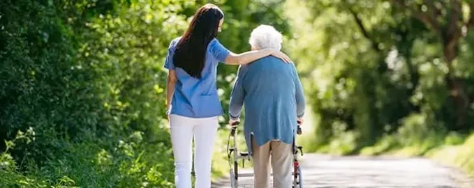 A caregiver in a blue uniform walks alongside an elderly person using a walker on a sunlit path surrounded by greenery. The caregiver gently places a supportive arm around the elders shoulders.