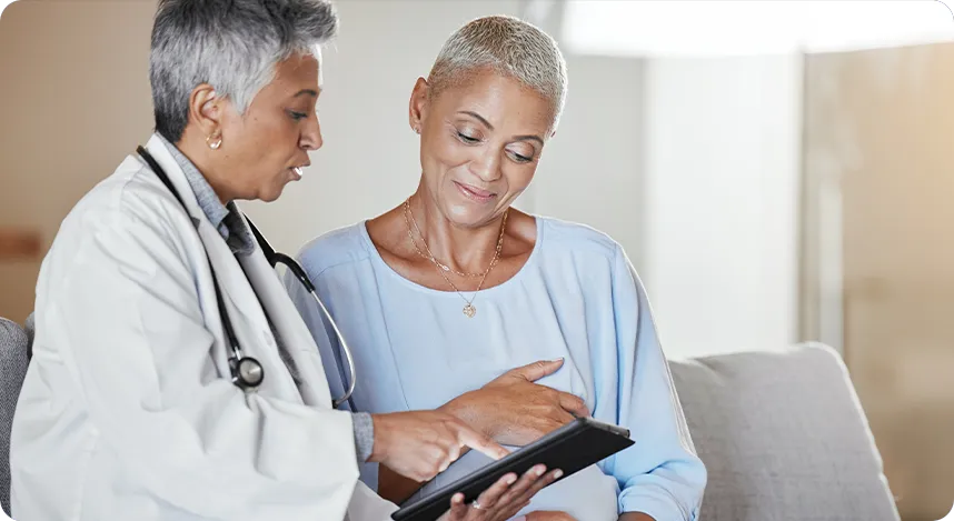 A doctor with a stethoscope shows a tablet to a patient. Both women are sitting and looking at the screen, engaged in discussion.