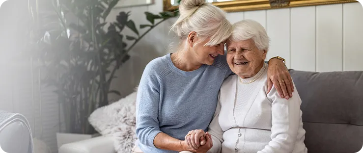 Two older women sitting on a couch, smiling and embracing. One has gray hair tied back and the other has short white hair.