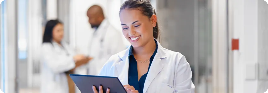 A woman in a lab coat smiles while using a tablet. Two other people in lab coats are blurred in the background.