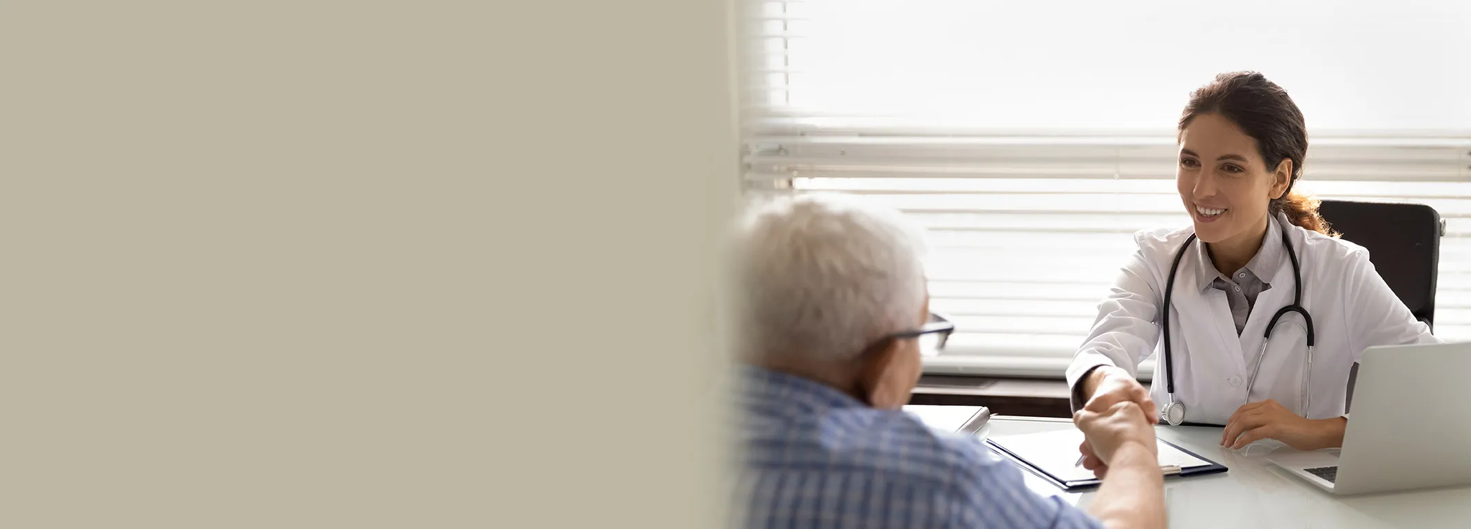 Doctor in a white coat smiling and shaking hands with an elderly person across a desk in a sunlit office.